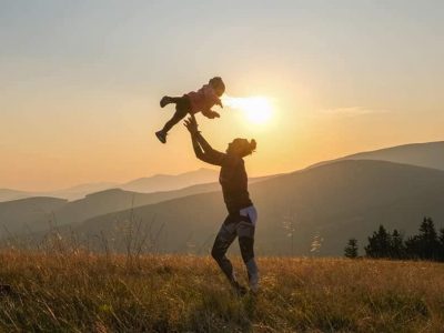 Dzikie dzieci, mama podrzuca córkę przy zachodzie słońca na polanie w górach - Podróże z dzieckiem